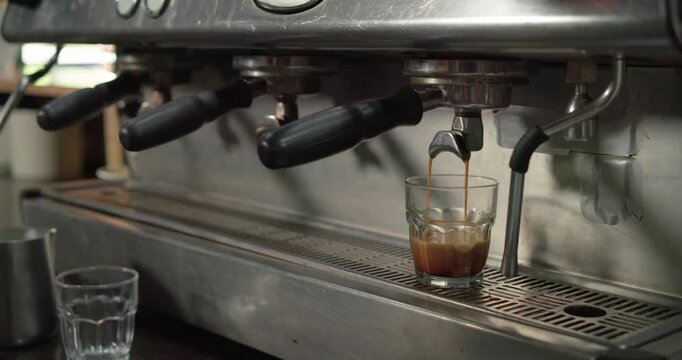 Machine spout is starting to release espresso into glass on drip tray, extracting crema for serving