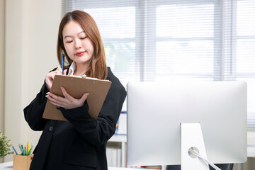 Young happy asian businesswoman holding clipboard and pen, working with documents and computer, sitting at desk in modern office ,business concept