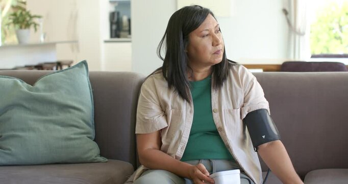 Asian mature woman sitting on living-room sofa checking BP with black cuff and monitor for health
