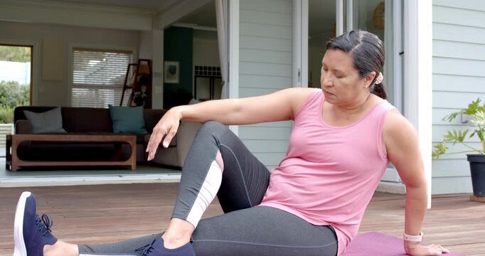 Mature woman in sportswear sitting left-hand behind on mat on deck, bending right knee for stretch