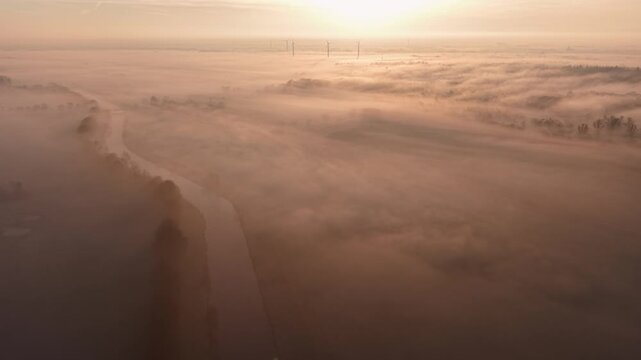 Aerial view of foggy canal at sunrise, flanked by trees and fields. Wind turbines silhouette the horizon as warm light diffuses through mist, evoking serenity, sustainability, and rural elegance.