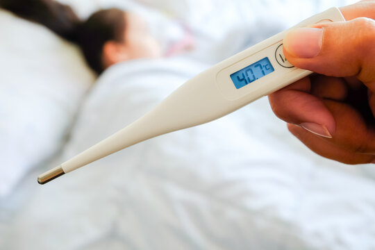 Close-up of a hand holding a digital thermometer showing a high fever of 40.7 degrees Celsius with a blurred sick person lying in bed, emergency medical and family healthcare concept.