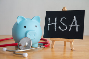 HSA hand writing on blackboard, piggy bank and stethoscope on wooden table. Health savings account (HSA), money management for medical treatment, health and life insurance, wellbeing and so on concept