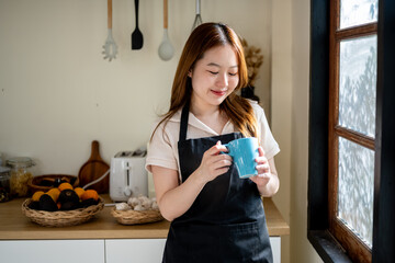 An Asian woman is happily drinking her morning coffee in the kitchen.