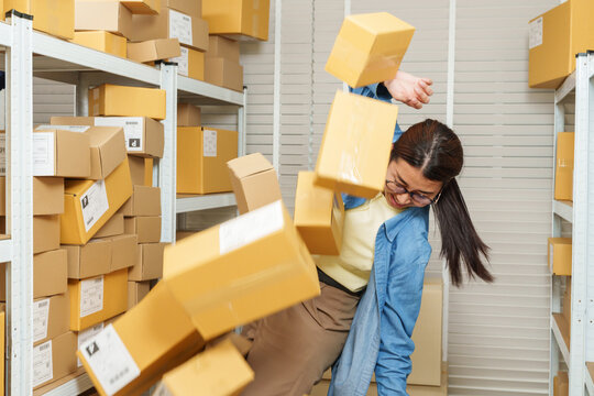 Asian adult woman small business owner losing balance while parcel boxes fall around warehouse aisle, ecommerce storage overload creating hazardous situation for online seller