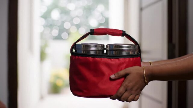 Hands Exchanging A Red Insulated Lunch Bag Containing Stainless Steel Tiffin Carriers With Food To Be Delivered To A Person Standing Near A Doorway With Blurred Greenery Outside Providing Natural