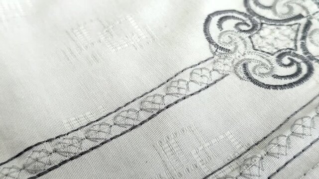 Close-up of grey embroidery on white fabric, featuring elegant scroll patterns and intricate stitch details on a traditional shirt