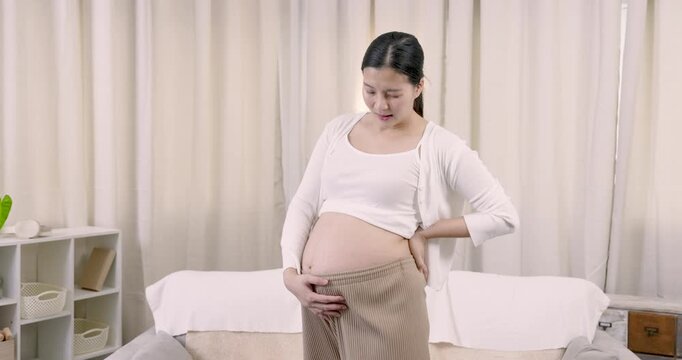 Asian pregnant woman standing beside bed while gently holding round belly expressing anticipation, care, and self-love during quiet third trimester maternity moment at home