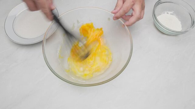 Hands whisking eggs in a glass bowl during cake preparation.