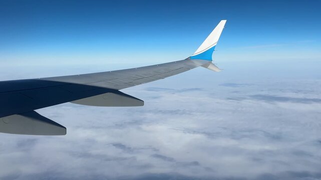 Passenger window view of a commercial airplane Boeing 737 Max wing above scattered clouds in a clear blue sky at cruising altitude, over clouds