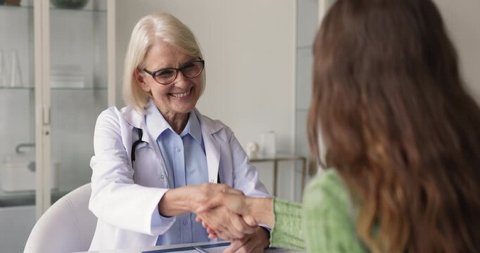 Middle age woman doctor provide professional medical assistance, gives health recommendations advice and medication prescription to female patient, shaking hands, finish consultation in clinic office