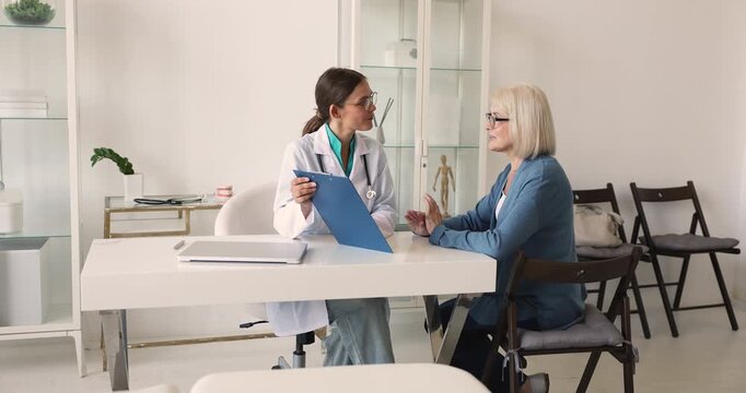 Doctor explains diagnosis or medical results on clipboard to middle age woman patient, review treatment plan, gives professional recommendations during her visit at hospital. Health care consultation