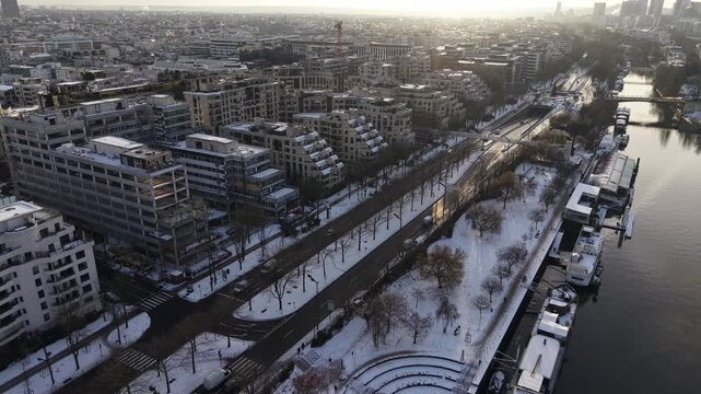 Cinematic aerial view of snow-covered riverfront promenade along Seine with modern buildings, winter trees, and bridges in Paris, France