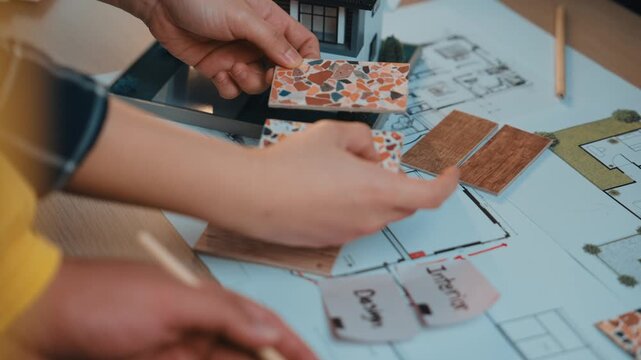 Two hands are seen holding colorful tiles and material samples over detailed interior design plans, illustrating a collaborative creative workspace focused on project development. SACTR