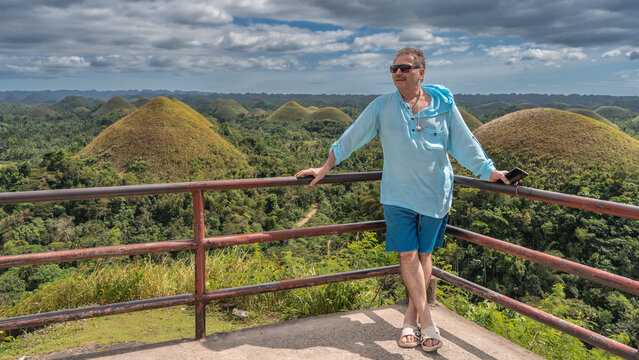 The man is standing on the observation deck, smiling, looking away, leaning on the railing. Rows of rounded conical hills in the valley. Blue sky, clouds. Philippines. Bohol. Chocolate Hills 