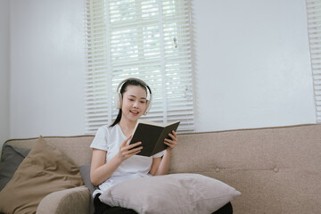 Obraz premium A young Asian woman relaxes on a sofa at home, wearing wireless headphones while reading a book, enjoying a calm moment of learning, entertainment, and modern digital lifestyle.