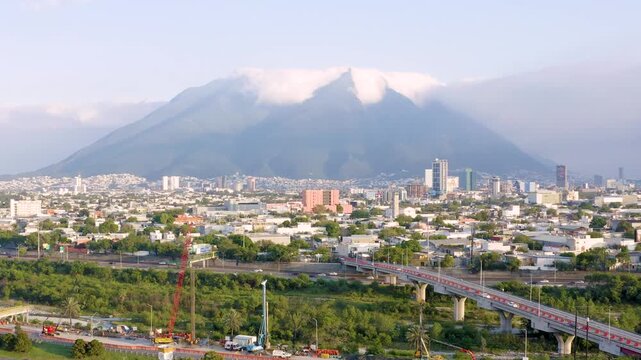 Cinematic forward-moving drone footage approaching Cerro de la Silla, Monterrey&rsquo;s iconic mountain, partially covered by soft white clouds. Below, traffic flows along a major highway.