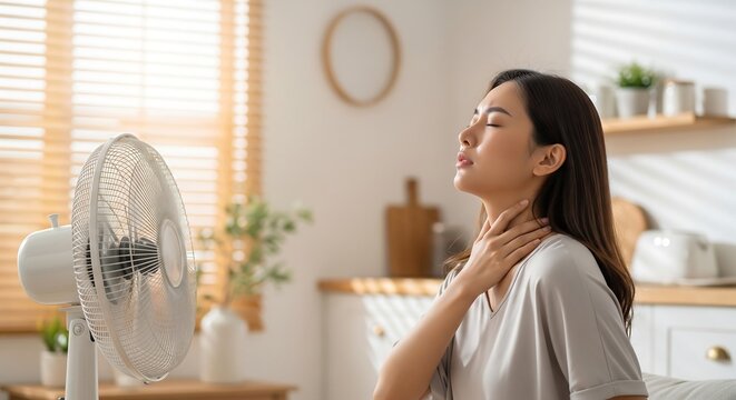 A young Asian woman feeling the heat and fanning herself with a portable electric fan in a sunlit room.