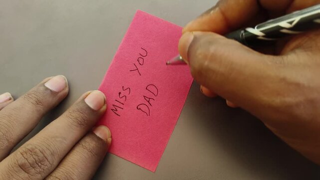 Indian or Asian person hand with pen writing miss you dad message or word on blank sticky note paper on a desk background. Handwritten reminder card expressing love, affection and bonding to father.