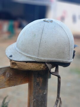 Close-up of a dusty vintage equestrian helmet resting on a wooden fence outdoors, horse riding safety equipment with visible wear and texture