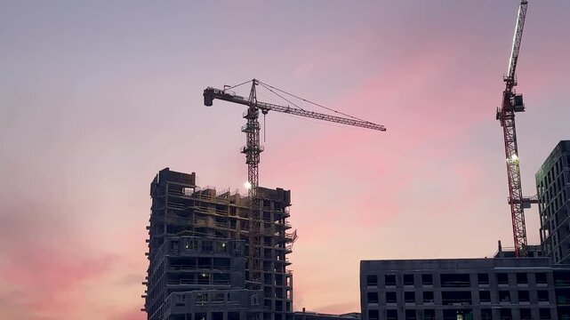 Construction site with towering cranes and multi-story residential buildings under development against a vibrant sunset or sunrise sky. Real estate investment and urban growth concept.
