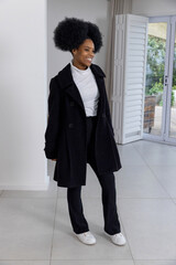 African American woman standing by shuttered sliding doors at home in long black coat, smiling