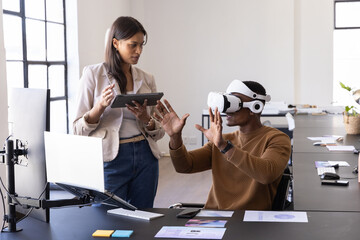 Diverse coworkers collaborating using vr headset and tablet at desk in modern office