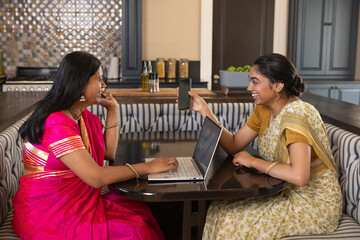 Indian mother and daughter wearing saris, sharing smartphone content and using laptop in kitchen © wavebreak3