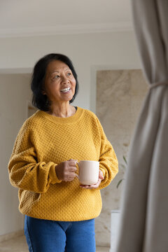 Senior Asian woman holding large ceramic mug smiling gazing right in living area wearing sweater