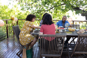 Diverse friends sitting around table on deck sharing chips, desserts on cake stand under bunting