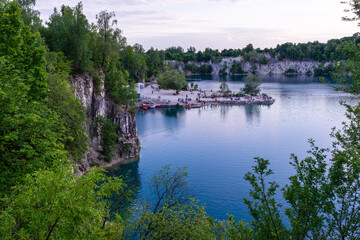 Fototapeta premium A panoramic view of Zakrzowek quarry lake near Krakow, showing steep cliffs, clear blue water, and a small recreational area with visitors along the shore. The scene captures summer leisure, nature to