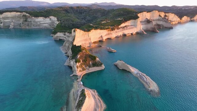 High-altitude aerial drone shot of Cape Drastis, Corfu, Greece, during a vibrant sunset. Wide-angle view of the entire coastal area, white cliffs, and sea stacks
