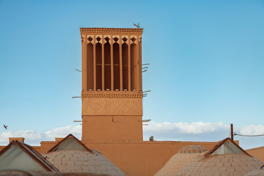 A wind catcher (badgir) rises above traditional domed rooftops, set against a bright blue sky. This ancient Persian ventilation system reflects the architectural ingenuity of Iran&rsquo;s desert regions.