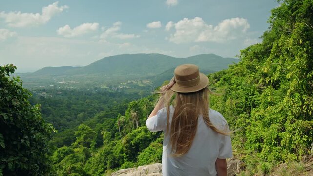 Young woman in straw hat standing on cliff viewpoint above Na Muang 2 Waterfall on Koh Samui, overlooking tropical jungle valley, green hills, and distant mountains with wide copy space sky