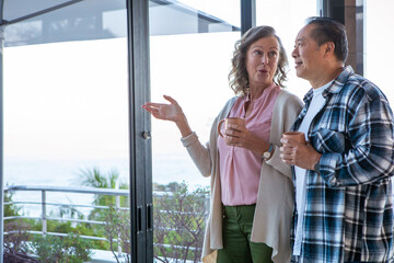 Diverse senior couple holding mugs by glass door in living room pointing toward ocean