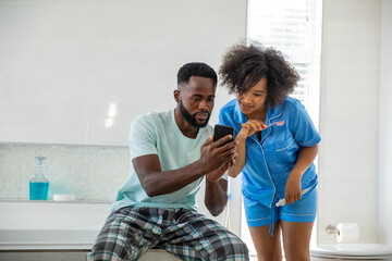 African american couple sitting on toilet lid checking smartphone, brushing teeth in bathroom