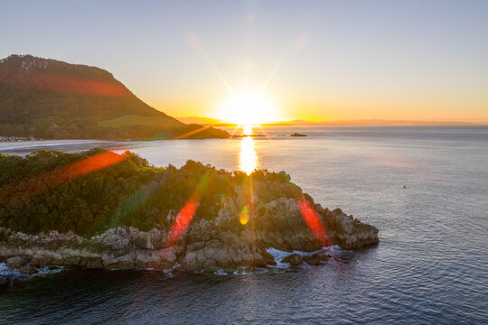 Aerial view of sun ablaze over the ocean, casting golden light on the craggy coast and verdant headland, Mount Maunganui, Bay of Plenty Region, New Zealand.