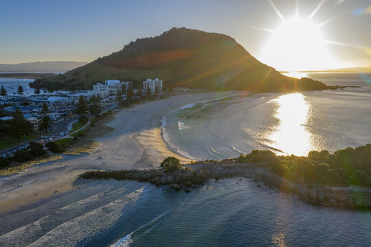 Aerial view of sun-kissed sands meet the tranquil turquoise waters at the base of a lush green mountain, Mount Maunganui, Bay of Plenty Region, New Zealand.