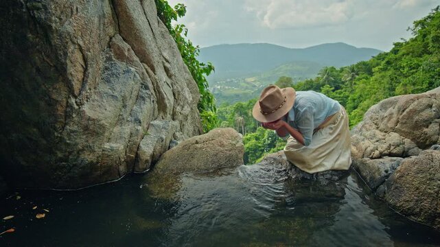 Middle-aged woman washing hands in clean natural rock pool at tropical waterfall overlook, peaceful travel scene with green valley, mountains, and calm outdoor lifestyle