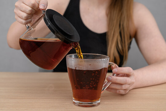 A woman pours hot tea from a teapot into a glass mug.