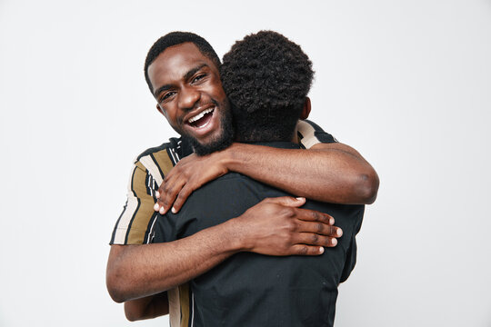 Two black men hugging each other with happy expressions on white background. African ethnicity, dark skin, friendship, connection, joy, casual clothing, positive emotions, studio portrait.