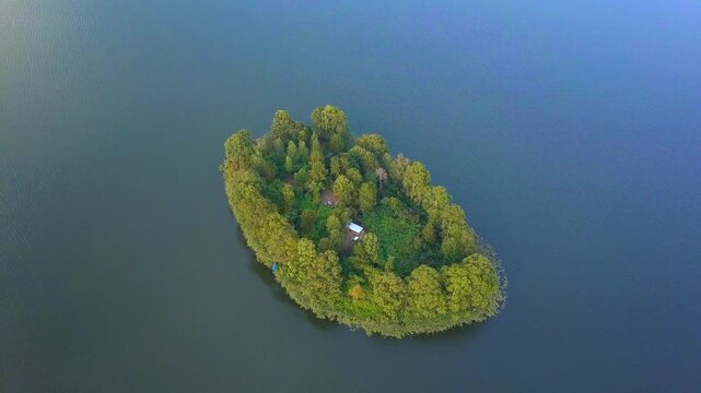 Lone small island with lush green trees and a visible house, surrounded by the calm, dark blue waters of Lake Bunyonyi, Uganda, from an aerial perspective