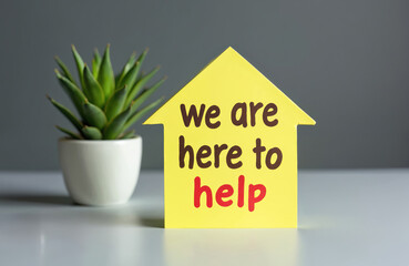 Fototapeta premium Yellow house shape with text we are here to help on gray background next to green plant in pot. Support, assistance, service, advice, offer.