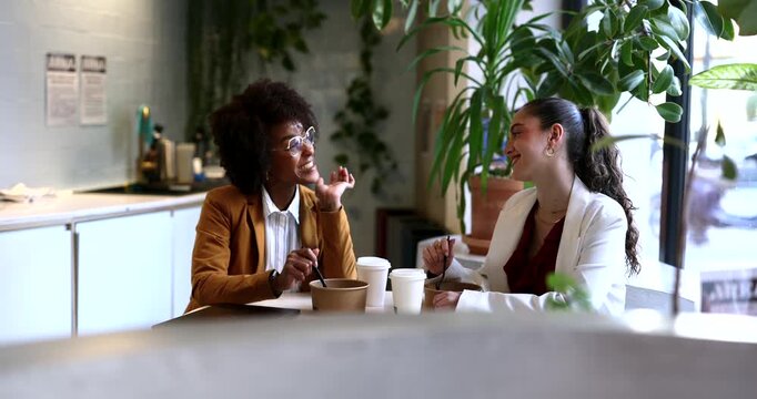 Two diverse businesswomen sitting at a table in a modern office breakroom, eating takeaway food from containers and engaging in a friendly, smiling conversation during their lunch break
