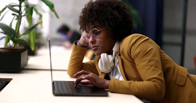 Overworked and exhausted black woman with curly hair and glasses leaning on her hand while struggling to stay focused and work on her laptop at the office, feeling sleepy and unmotivated