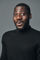 Obraz premium African man with dark skin wearing black turtleneck sweater in creative conceptual visual style looking surprised with open mouth and expressive face on gray background studio portrait.