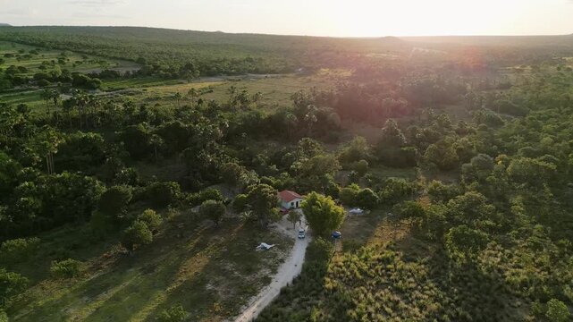 Aerial drone view captures a rural homestead fazenda in the Cerrado biome of Goi&aacute;s, Brazil, highlighting a single house, unpaved access road, surrounding native vegetation, and warm sunset light