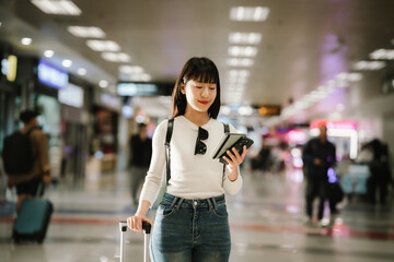 Obraz premium A smiling young Asian woman walks through an airport terminal holding a smartphone and rolling suitcase, representing modern travel, confidence, mobility, and a contemporary lifestyle.