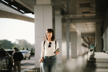 Fototapeta premium A young Asian woman with a backpack and rolling suitcase smiles while checking her smartphone at a modern transit station, capturing the concept of travel, mobility, and urban lifestyle.