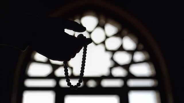 A silhouette of a hand holding prayer beads against a geometrically patterned window, evoking spirituality.